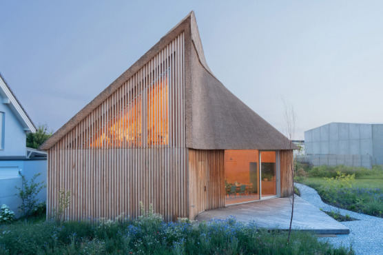 Ein modernes schilfgedecktes Einfamilienhaus aus Holz. Innen ist warmes Licht zu sehen.